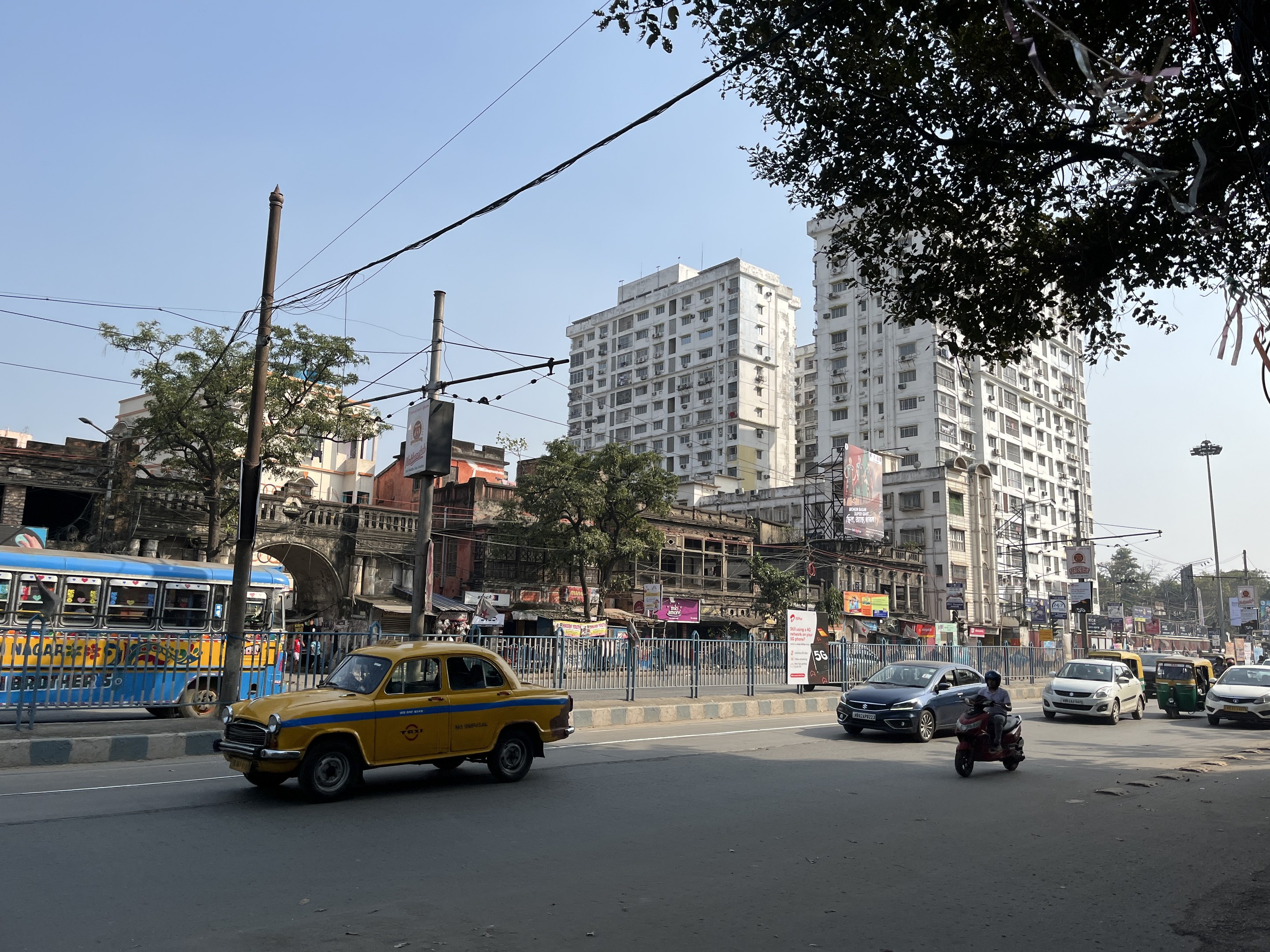 Gelbes Taxi fährt auf einer belebten Straße in Mumbai, mit hohen Gebäuden, Bäumen und Strommasten im Hintergrund.