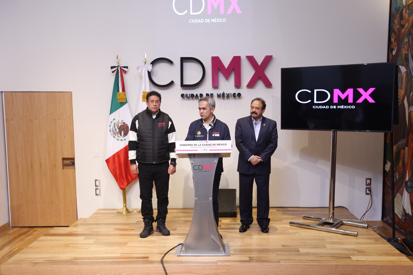 Drei Männer stehen an einem Podium mit einem Mikrofon, flankiert von Flaggen, einer Tür und einer textbedeckten Wand, während sie auf einer Pressekonferenz in Mexiko vor einem Fernseher und einem Banner sprechen.