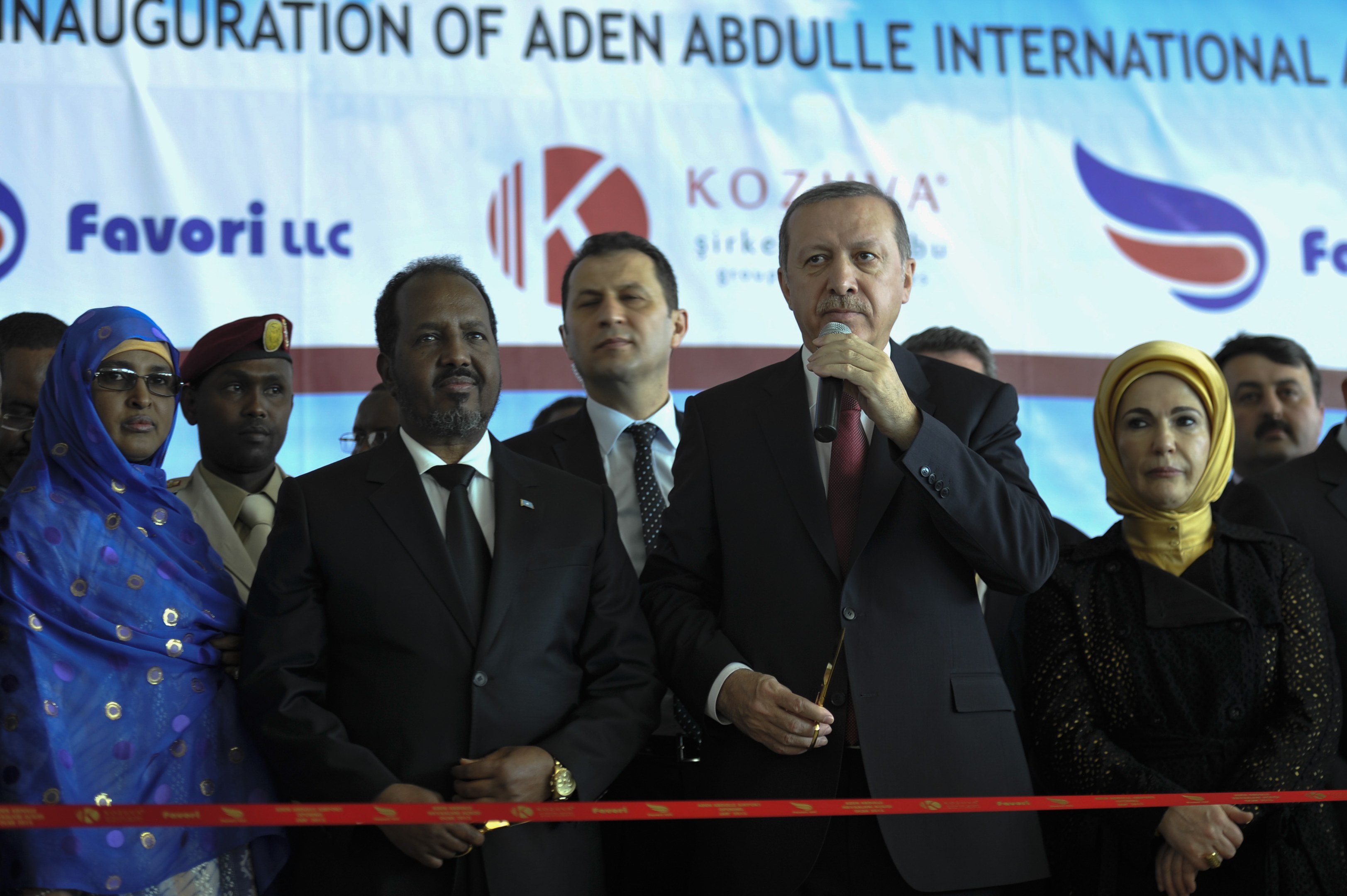 Gruppe von Menschen vor einer roten Schleife mit einem Mann mit Mikrofon, dahinter eine Tafel mit der Aufschrift 'Eröffnung des Aden Abdulle International Airport'.