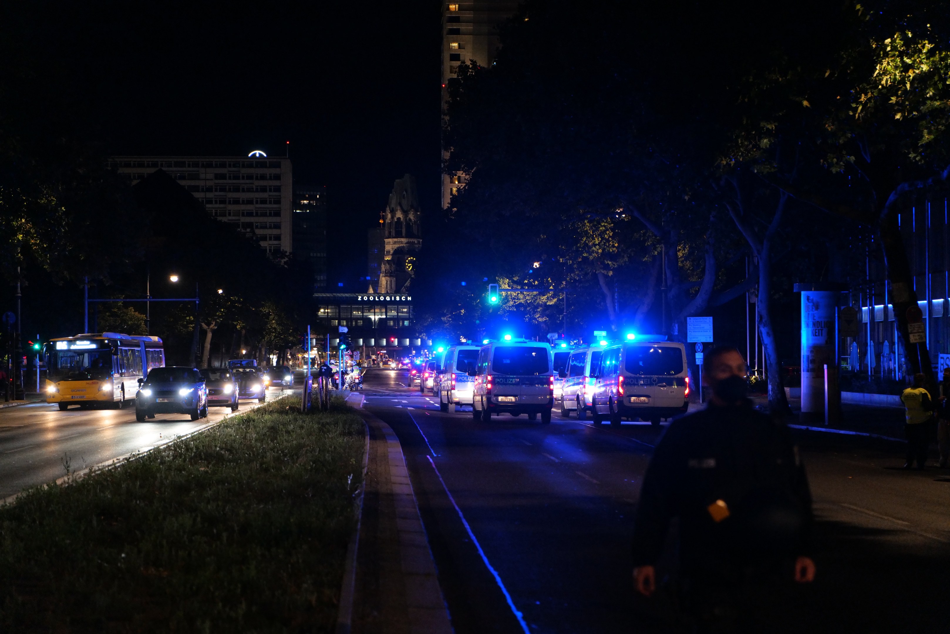 Stadtstraße bei Nacht mit Polizeiwagen und Bussen, die fahren, Menschen auf dem Bürgersteig, Gras, Bäume, Gebäude und beleuchtete Laternen.