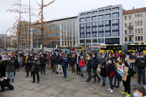 Eine große Gruppe von Menschen, einige mit Masken und Schildern, steht vor einem Gebäude während einer Protestaktion in Berlin, mit Fahrzeugen, Bäumen, Laternenmasten und Gebäuden mit Fenstern im Hintergrund und dem Himmel darüber.