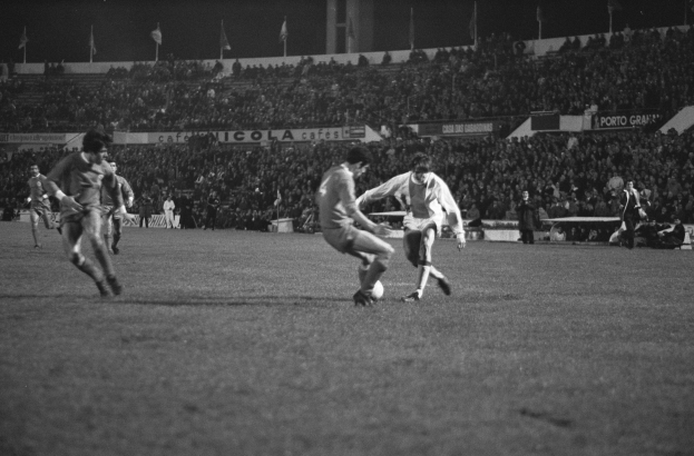 Schwarzes und weißes Bild von Männern, die Fußball auf einem Feld mit Zuschauern in den Stadionrängen und Fahnen im Hintergrund spielen.