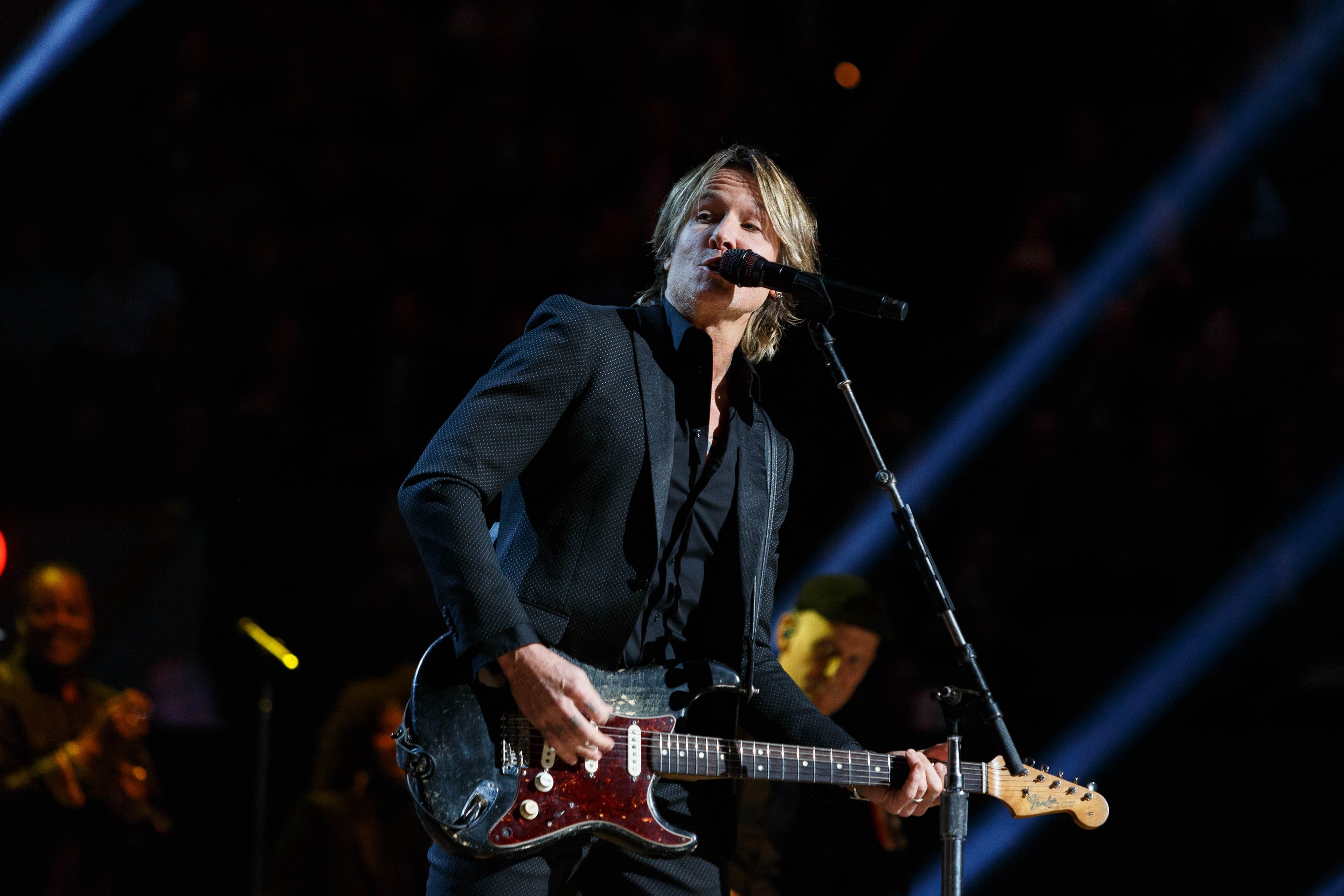 Keith Urban performs on stage playing an electric guitar with a microphone in front of him, accompanied by a few musicians in the background during a dimly lit concert at the Joint in Las Vegas.