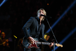 Keith Urban performs on stage playing an electric guitar with a microphone in front of him, accompanied by a few musicians in the background during a dimly lit concert at the Joint in Las Vegas.