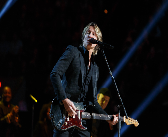 Keith Urban performs on stage playing an electric guitar with a microphone in front of him, accompanied by a few musicians in the background during a dimly lit concert at the Joint in Las Vegas.