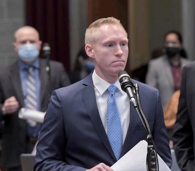 Ein Mann in Anzug und Krawatte spricht an einem Rednerpult mit Papieren, während er ein Mikrofon ansieht, während andere in der Nähe stehen.