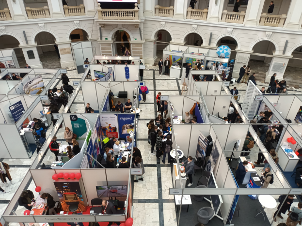 Große Menschenmenge auf einer internationalen Fachmesse mit Ständen, Bannern, Tischen, Stühlen, Luftballons und einem Gebäude mit Bögen, Säulen, Geländern und einem Bildschirm im Hintergrund.