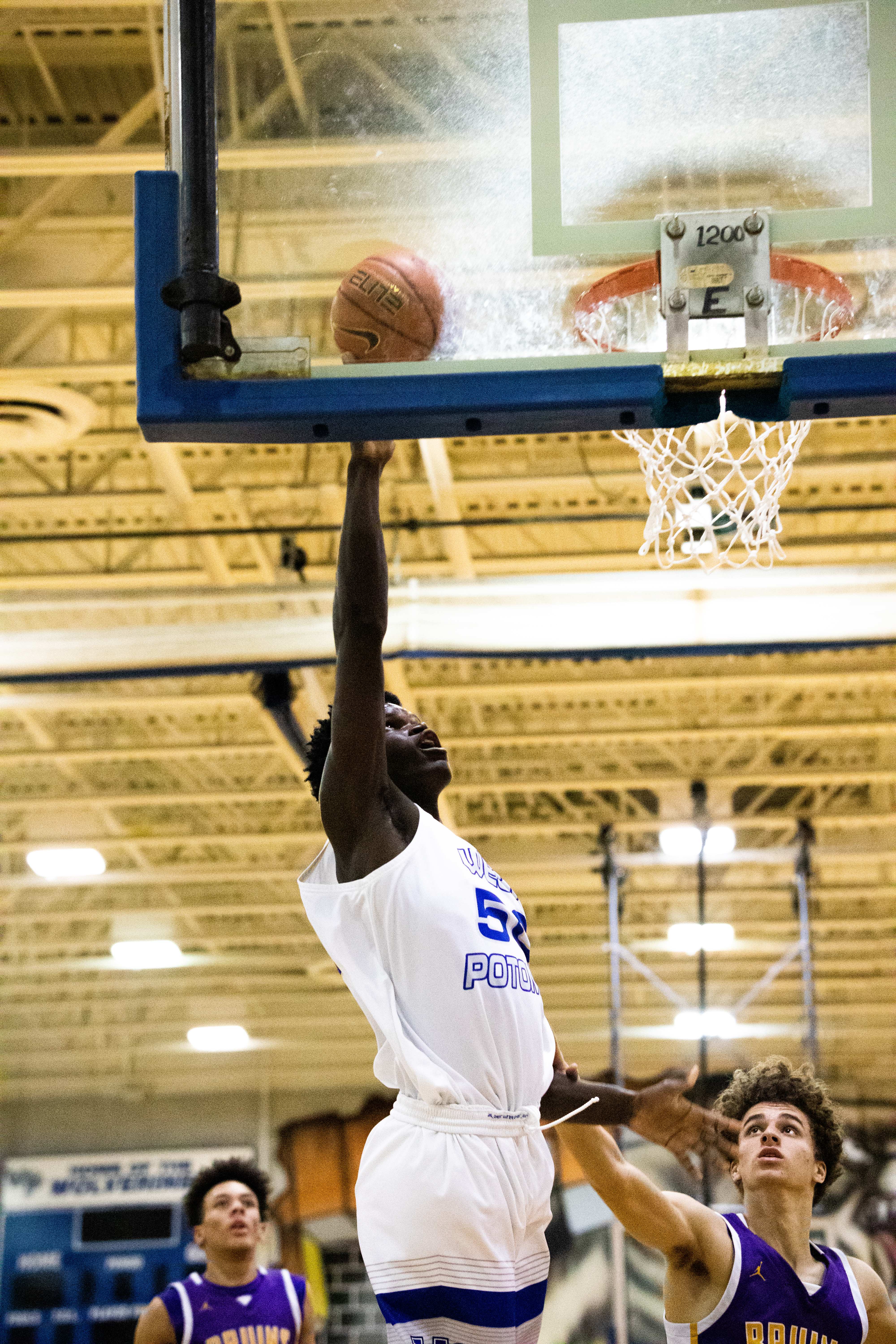 Ein Mann in weiß-blauer Uniform wirft einen Basketball in einer Turnhalle, an der ein Basketballkorb an einem Pfosten befestigt ist und Lampen an der Decke leuchten, während im Hintergrund andere Menschen zu sehen sind.