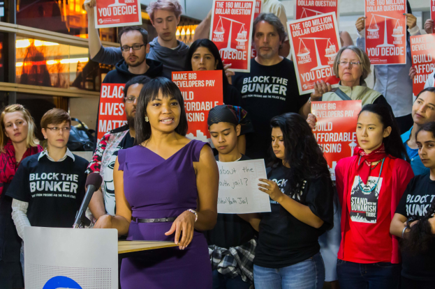 Frau mit Mikrofon an einem Rednerpult, spricht zu einer Menge mit Schildern, identifiziert als Black Lives Matter-Aktivistin.
