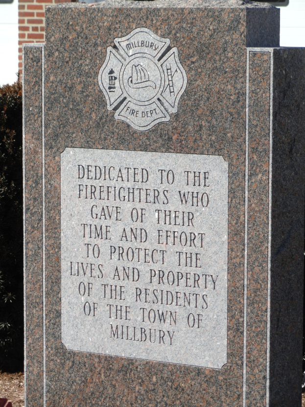 Denkmalstein mit einer Gedenktafel für Feuerwehrleute in Millbury, vor einem Backsteingebäude mit umliegenden Pflanzen und Bäumen gesetzt.