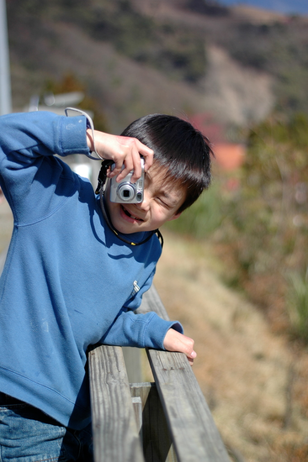 Ein Mann in einem blauen Hemd und Jeans hält eine Kamera vor der Stadt, mit Bäumen und Himmel im Hintergrund.