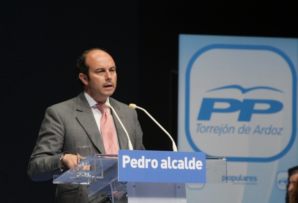 Mann in Anzug spricht an einem Pult mit Mäusen und einem Glas darauf, mit schwarzem Hintergrund und einer Tafel rechts.