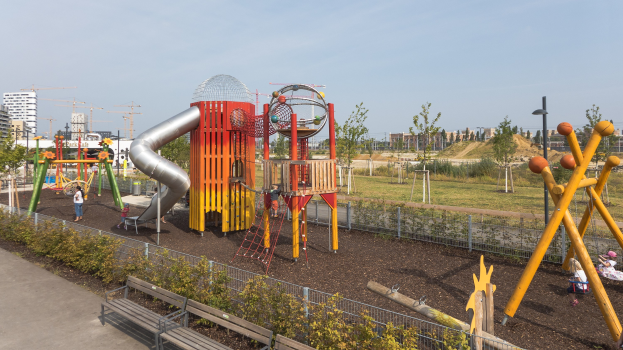 Spielplatz mit farbenfroher Ausstattung, einschließlich einer Rutsche und Schaukeln, umgeben von Pflanzen, einem Zaun, Bänken und einer Straße, mit Bäumen, Gebäuden und Kränen im Hintergrund unter einem sichtbaren Himmel.