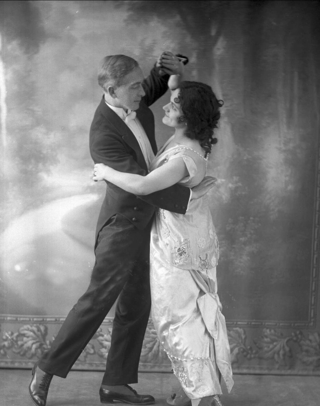 Schwarzes und weißes Foto eines Mannes in einem Anzug und einer Frau, die Tango tanzen, elegant über einen Boden mit einem gemusterten Hintergrund gleiten.