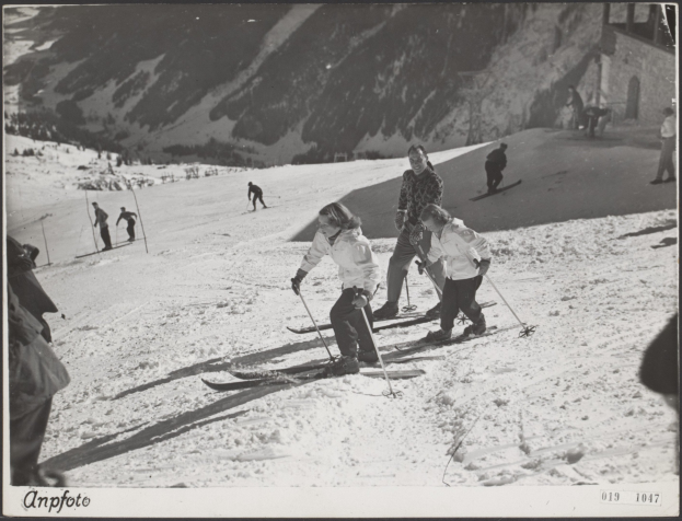 Gruppe von Menschen, die eine schneebedeckte Piste mit Skistöcken hinunterfahren, mit Hügeln und einem Gebäude im Hintergrund und Text unten.