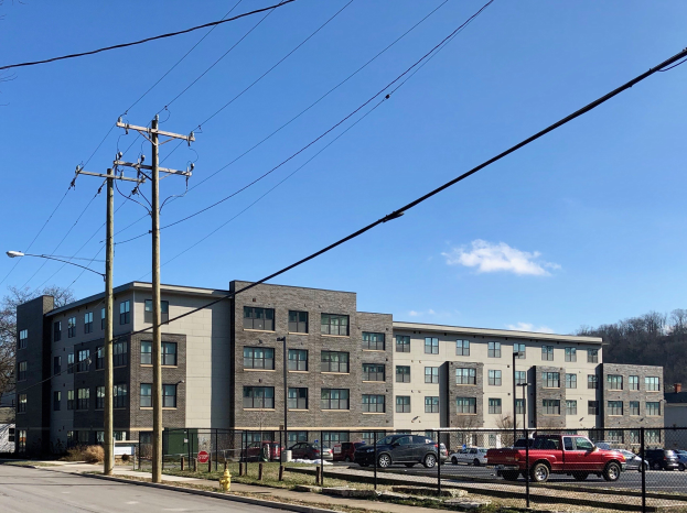 Ein großes Wohnhaus an einer Straßenecke mit Strommasten, Kabeln, Straßenlaternen, Fahrzeugen, einem Hydranten, einem Zaun, Bäumen und einem bewölkten Himmel.