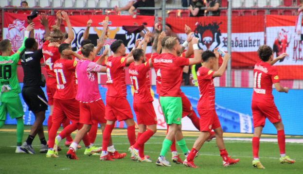Gruppe von Fußballspielern, die ein Tor feiern, auf einem Feld mit Flaschen haltenden Spielern und klatschenden Spielern, einem Zaun, Bannern, Metallstangen, Stühlen und einer Decke mit Deckenleuchten im Hintergrund.