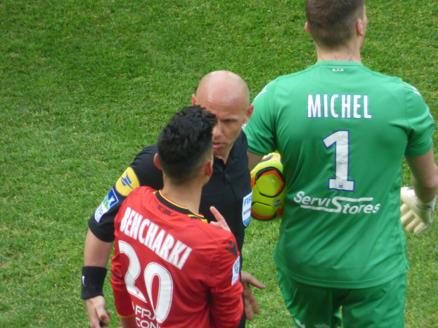 Schiedsrichter spricht mit zwei Fußballspielern auf einem grünen Rasen, ein Spieler hält einen Fußball.