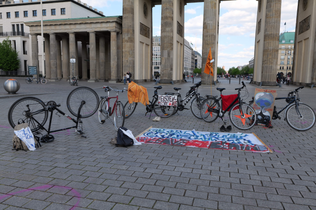 Eine Gruppe von Fahrr├Ądern, die vor dem Brandenburgertor mit Taschen und einer Fahne in der N├Ąhe geparkt sind, vor einer Kulisse aus Geb├Ąuden, B├Ąumen, Masten, Flaggen und einem klaren blauen Himmel.