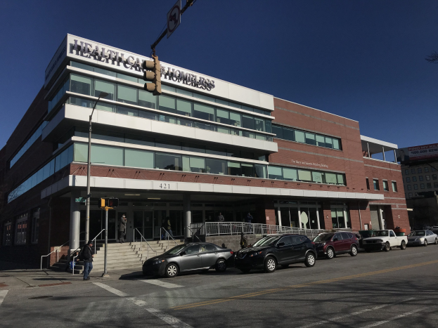 Großes Gebäude als New York City Health Care for the Homeless Center identifiziert mit Autos davor, umgeben von Straßeninfrastruktur und Passanten unter einem klaren blauen Himmel.