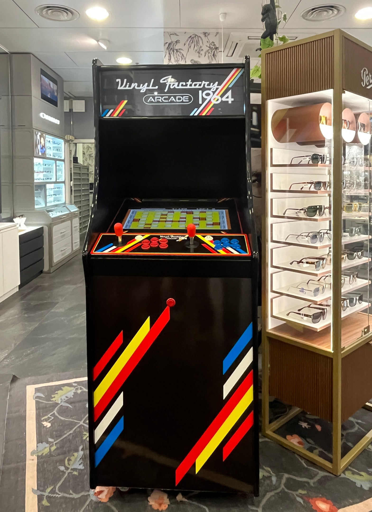 Ein Geschäft mit einem zentralen Retro-Arcade-Automaten, einer Glasvitrine auf der rechten Seite und Retro-Beleuchtung mit Schränken, Fernseher und anderen Gegenständen im Hintergrund.