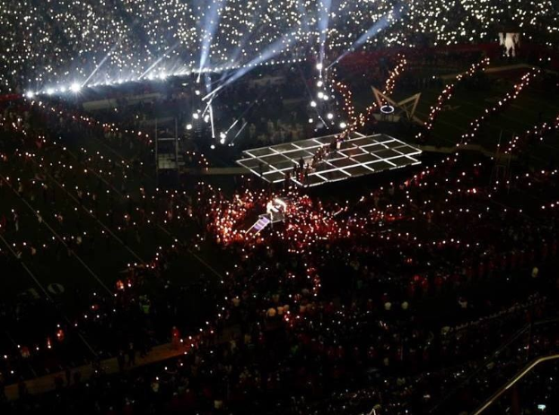 Eine große Menschenmenge in einem hell erleuchteten Stadion bei Nacht, mit einer Person, die auf einer zentralen Bühne während der Olympischen Schlussfeier performs.