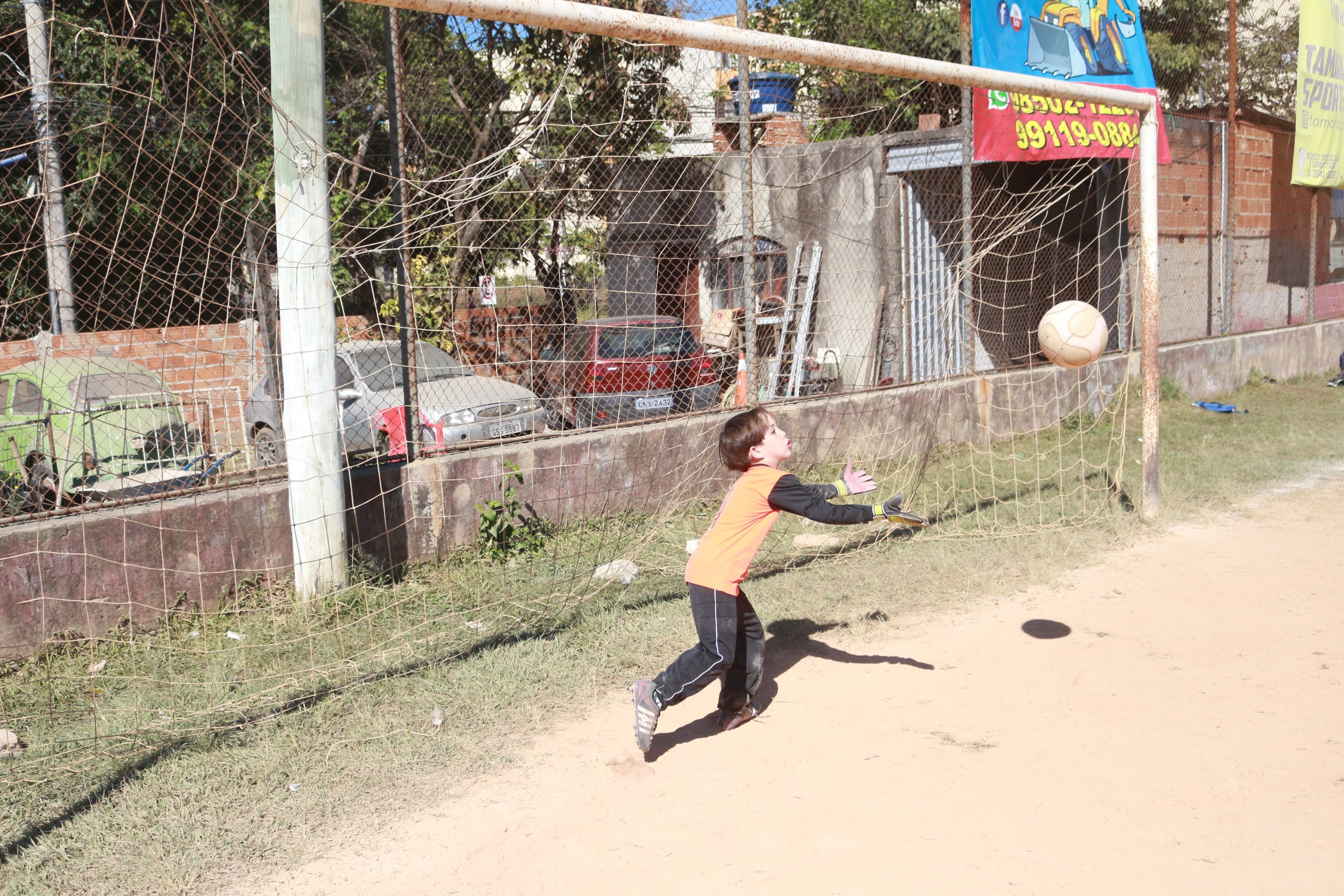 Ein junger Junge, der einen Füüball in ein Tor schießtzt, mit Pfosten, Bannern, Gebäuden, Bäumen, Fahrzeugen und einem klaren blauen Himmel im Hintergrund.