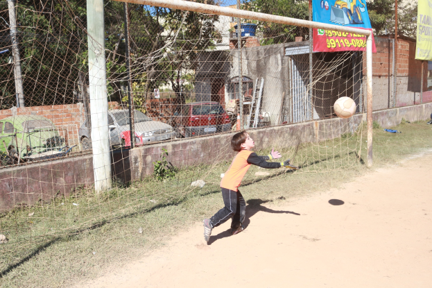 Ein junger Junge, der einen Füüball in ein Tor schießtzt, mit Pfosten, Bannern, Gebäuden, Bäumen, Fahrzeugen und einem klaren blauen Himmel im Hintergrund.
