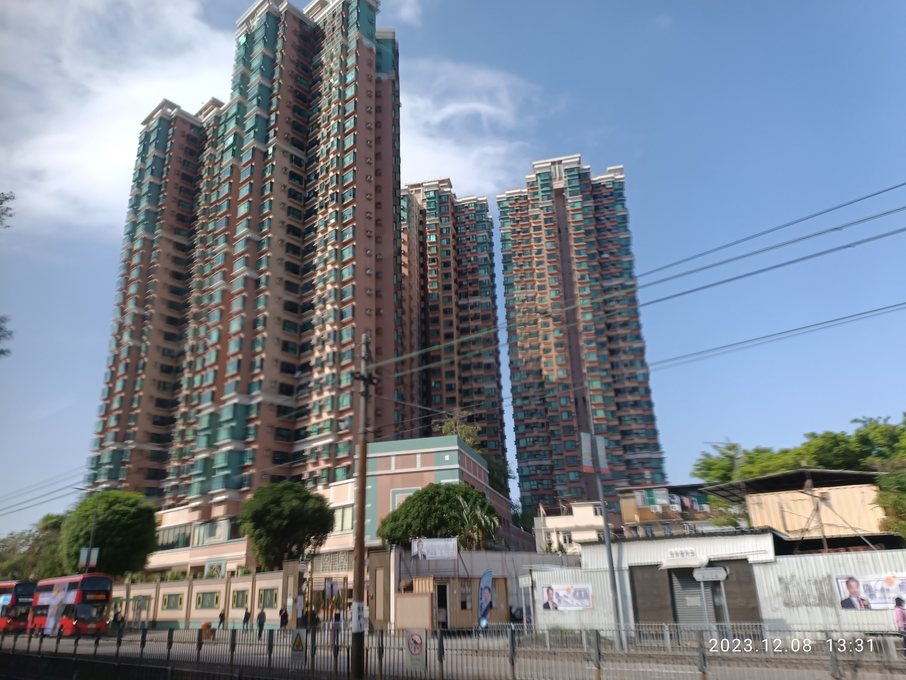 Lebendige Stadtstraße mit hohen Gebäuden, Fahrzeugen, einem Zaun, Pfählen, Schildern, Bäumen und Menschen in Mumbai unter einem bewölkten Himmel mit einem Wasserzeichen.