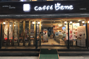 Ein Café-Gebäude in Seoul, Südkorea, mit Glaswänden und -türen, beleuchtet und geschmückt mit Text- und Bildtafeln, enthaltend Tische, Stühle und eine Wanduhr, neben einer Straße gelegen.