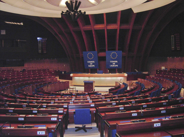 Das Europäische Parlament in Brüssel, Belgien, mit einem halbrunden Sitzplatzanordnung, zentralem Podium und an der Wand montierten Texttafeln unter einer beleuchteten Decke.