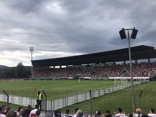 Große Zuschauermenge bei einem Fußballspiel in einem bewölkten, offenen Stadion mit beleuchtetem Feld, eingezäuntem Rand, Laternenmasten und fernen Bäumen und Hügeln.
