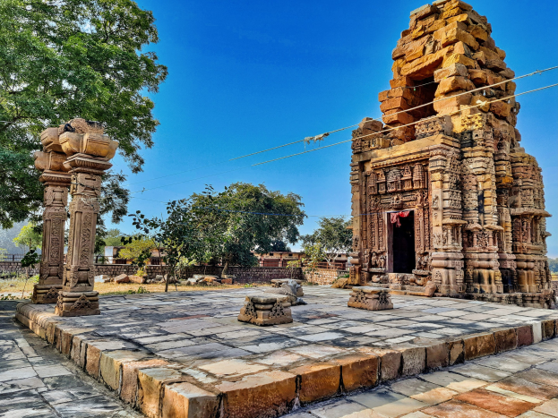 Ruine eines Hindu-Tempels in Hampi, Karnataka, Indien, mit verstreuten Säulen, Felsen, Bäumen und einer teilweisen Wand, mit dem Himmel im Hintergrund.