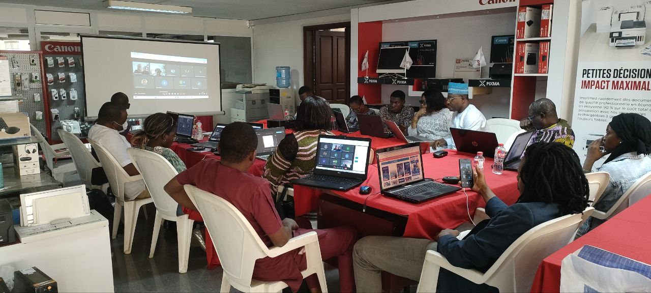 Gruppe von Menschen an einem Tisch mit Laptops und anderen Gegenständen während eines digitalen Marketing-Workshops, mit einem Banner, einem Display und einer Tür im Hintergrund.