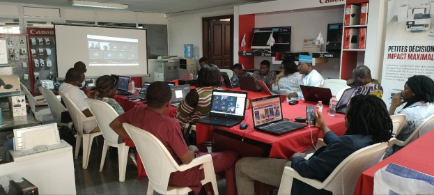 Gruppe von Menschen an einem Tisch mit Laptops und anderen Gegenständen während eines digitalen Marketing-Workshops, mit einem Banner, einem Display und einer Tür im Hintergrund.