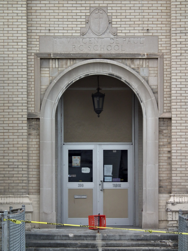 Ein Eingang eines Backsteingebäudes mit gelbem Absperrband, Treppen mit Maschendrahtzaun auf beiden Seiten, eine Tür mit daran geklebten Papieren, eine Deckenlampe und eine Wandinschrift, die wahrscheinlich den Namen der Schule angibt.