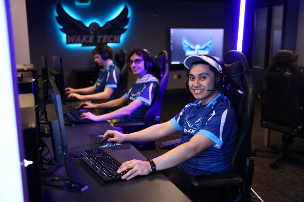 Eine Gruppe von Menschen, die an Computern sitzen und Headsets tragen, lächeln und mit Monitoren, Tastaturen und anderen Gegenständen auf dem Schreibtisch, vor einer Wand mit Logo und Text und Stühlen rechts.