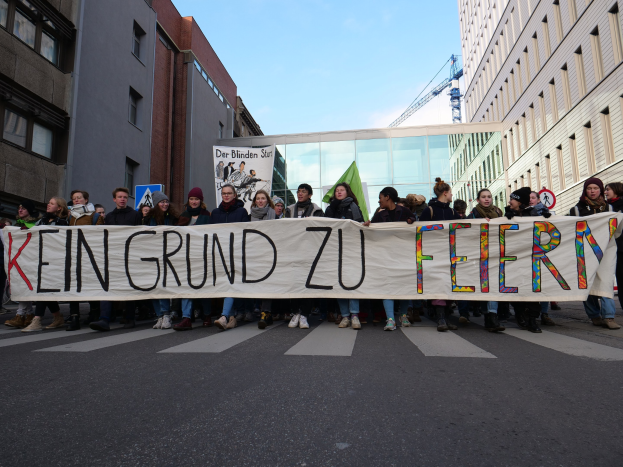 Demonstranten halten ein Transparent mit der Aufschrift 'Kein Grund zur Feier' gegen deutsche Sparmaßnahmen, mit Gebäuden, einem Kran und einem bewölkten Himmel im Hintergrund.