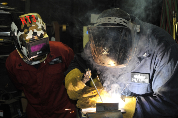 Zwei Schweißer in Helmen bei der Arbeit an einem Metallteil in einer Werkstatt, mit Rauch vom Schweißen und verstreuten Werkzeugen drumherum.