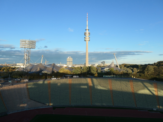 Das Olympische Stadion in Berlin, Deutschland, mit dem Fernsehturm (Fernsehturm) im Hintergrund, umgeben von Bäumen, Gebäuden und beleuchteten Bereichen unter einem bewölkten Himmel.
