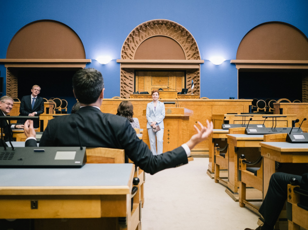 Eine Gruppe von Menschen sitzt an Schreibtischen in einem Gerichtssaal, eine Frau steht in der Mitte, Laptops und Mikrofone auf den Schreibtischen und beleuchtete Wände, was eine angespannte Atmosphäre vermuten lässt.