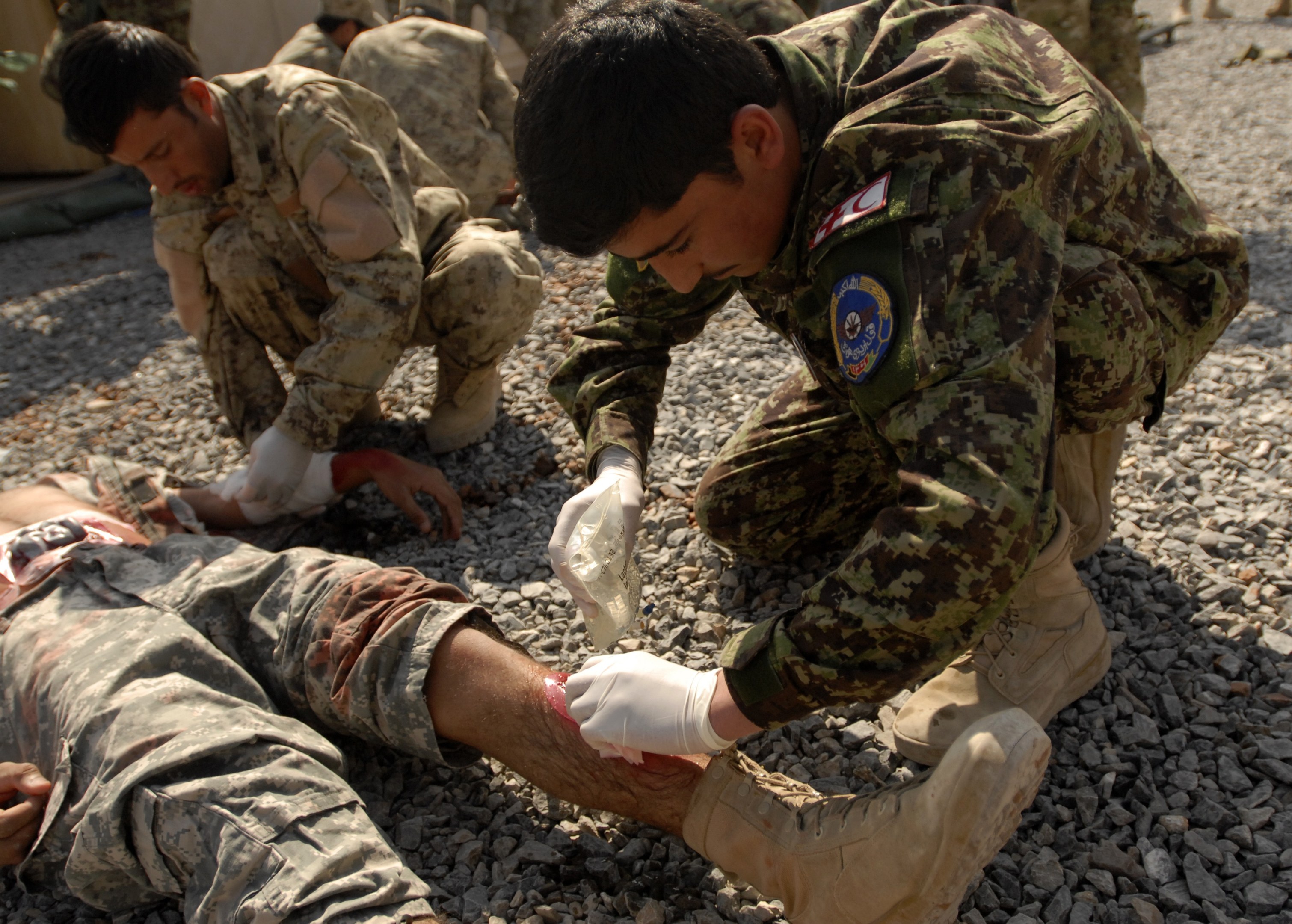 Gruppe von Männern in Militäruniformen kniend auf dem Boden, einer liegt auf Steinen mit einem verbundenen Bein, andere im Hintergrund neben einem Zelt.