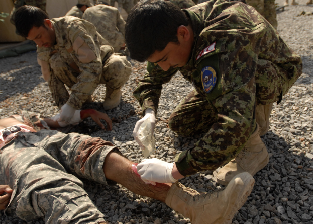 Gruppe von Männern in Militäruniformen kniend auf dem Boden, einer liegt auf Steinen mit einem verbundenen Bein, andere im Hintergrund neben einem Zelt.